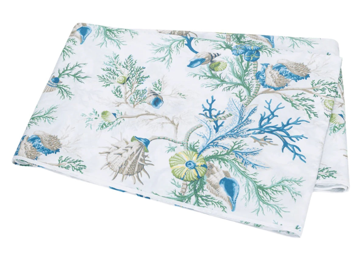 Immerse yourself in the Del Tesoro Bedding Collection by Matouk, featuring folded fabric adorned with a marine-themed print of blue and green shells, seaweed, and coral on a crisp white background—capturing an oceanic fantasy teeming with graceful maritime creatures.