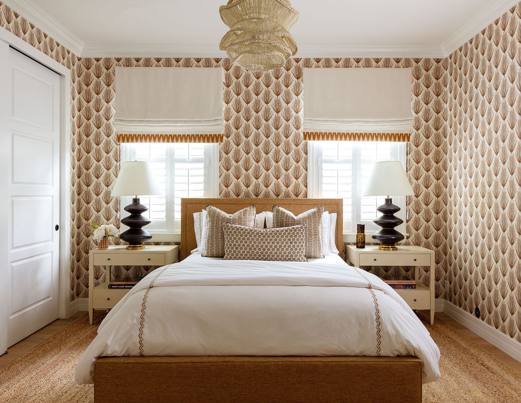 image of bedroom with brown palm leaf print wallpaper and coordinating white and brown tropical print bed pillows, natural jute rug, black ceramic lamps and white roman shades