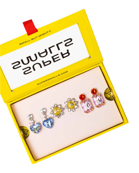 The *Super Smalls Dinner & A Movie Clip-On Earring Set* comes in a yellow box from *Super Smalls*. It contains three pairs of high-wattage sparkle clip-on earrings: blue heart-shaped, yellow flower-shaped, and pink rectangular-shaped. Inside the lid is a message that reads "SMALL BUT MIGHTY.