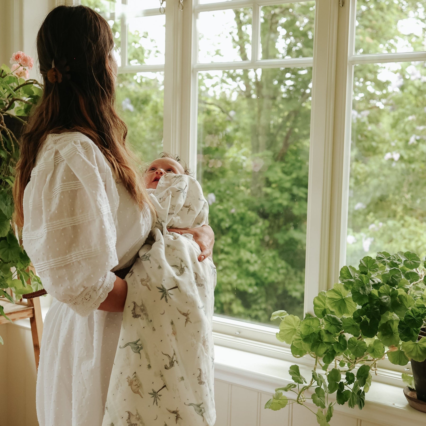 A woman in a white dress holds her baby wrapped in a Gooselings Dino Dreams Swaddle, standing by a large window with green plants on the windowsill.