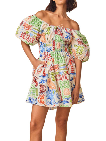 A woman wearing a Hunter Bell NYC Drew Dress, a colorful off-shoulder mini dress featuring a vibrant print with various motifs and a scoop neckline.