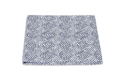 A folded fabric square from the Matouk Duma Diamond Bedding Collection, featuring a blue and white harlequin-inspired diamond motif, is displayed against a plain white background.