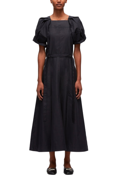 A woman stands facing forward wearing a black 3.1 Phillip Lim Collapsed Bloom Short Sleeve Belted Dress with puff sleeves and a cinched waist, paired with black shoes.