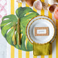 A tropical table features a white plate on a Hester & Cook Die Cut Monstera Leaf Placemat, gold cutlery, a yellow-striped tablecloth, and a "jessica" name card, all surrounded by seashells.
