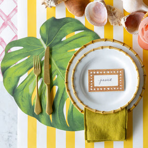 A tropical table features a white plate on a Hester & Cook Die Cut Monstera Leaf Placemat, gold cutlery, a yellow-striped tablecloth, and a "jessica" name card, all surrounded by seashells.