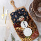 A festive charcuterie board featuring a cheese snowman, berries, blueberries, crackers, nuts, chocolate, salami, and popcorn is artfully arranged on the Be Home Acacia Wood Rectangular Board with Handle Collection.