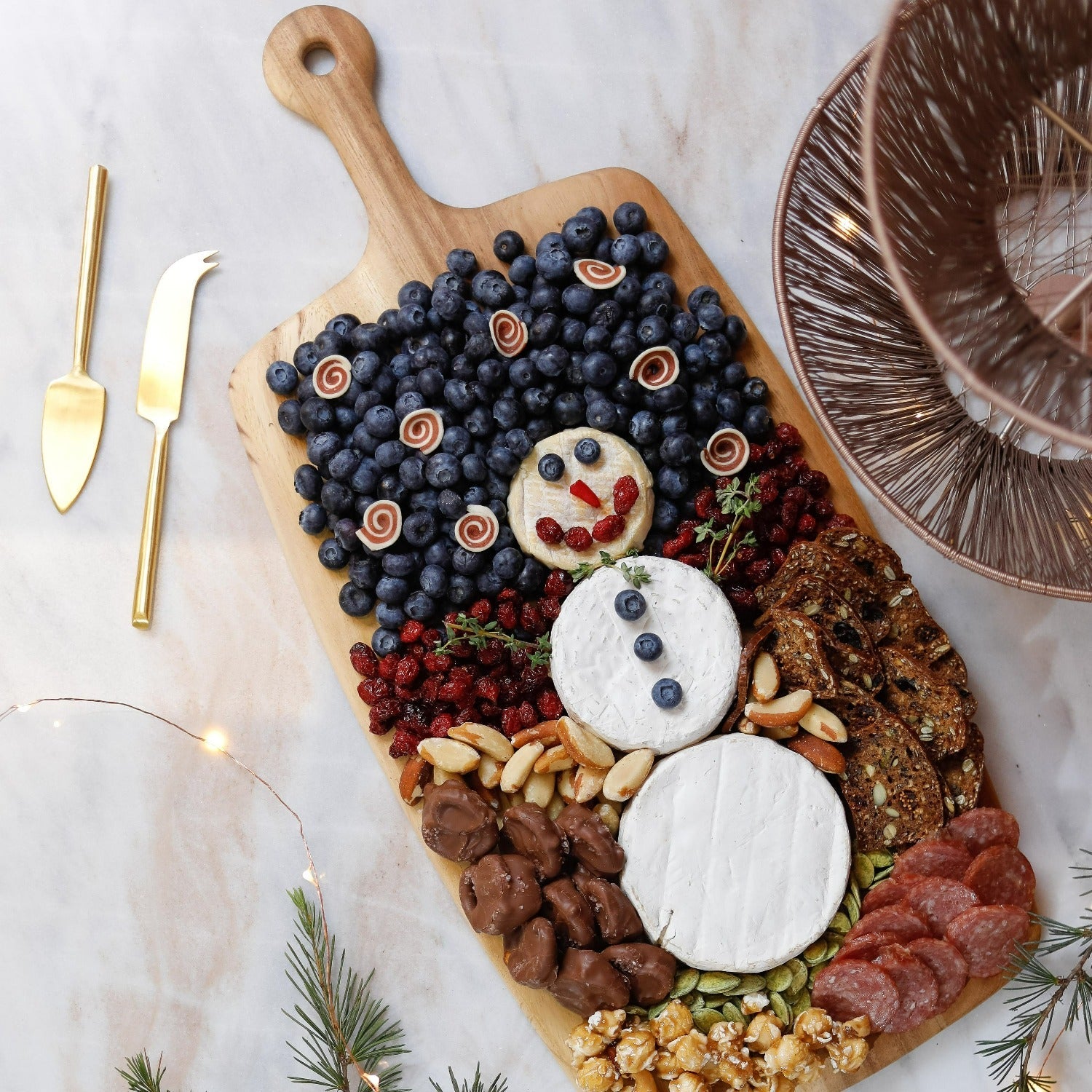 A festive charcuterie board featuring a cheese snowman, berries, blueberries, crackers, nuts, chocolate, salami, and popcorn is artfully arranged on the Be Home Acacia Wood Rectangular Board with Handle Collection.