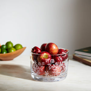 The Simon Pearce Berry Bowl, made of clear lead-free glass with a white floral pattern, holds small red apples on marble; limes and books appear in the background, creating a charming centerpiece.
