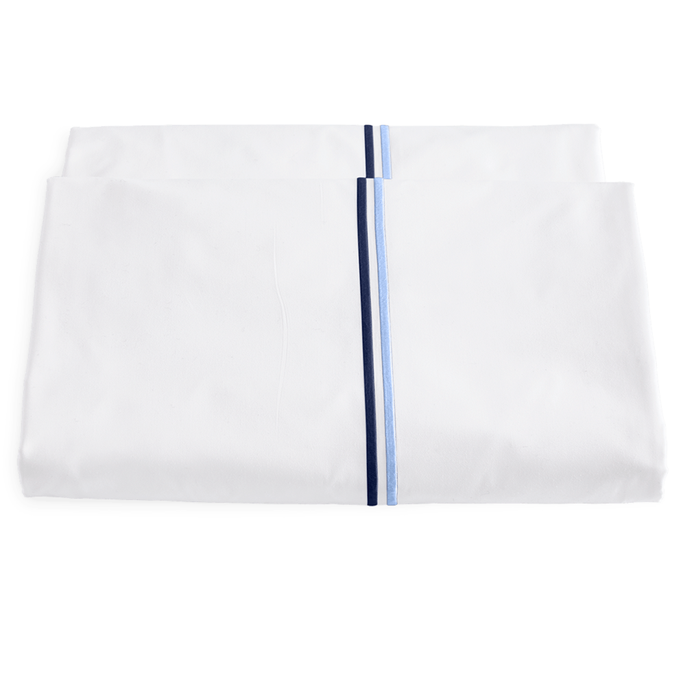 Two folded white sheets from the Matouk Essex Bedding Collection, Navy, featuring parallel blue and navy satin stitch embroidery near the edge, inspired by European hotel bedding, displayed on a plain white background.