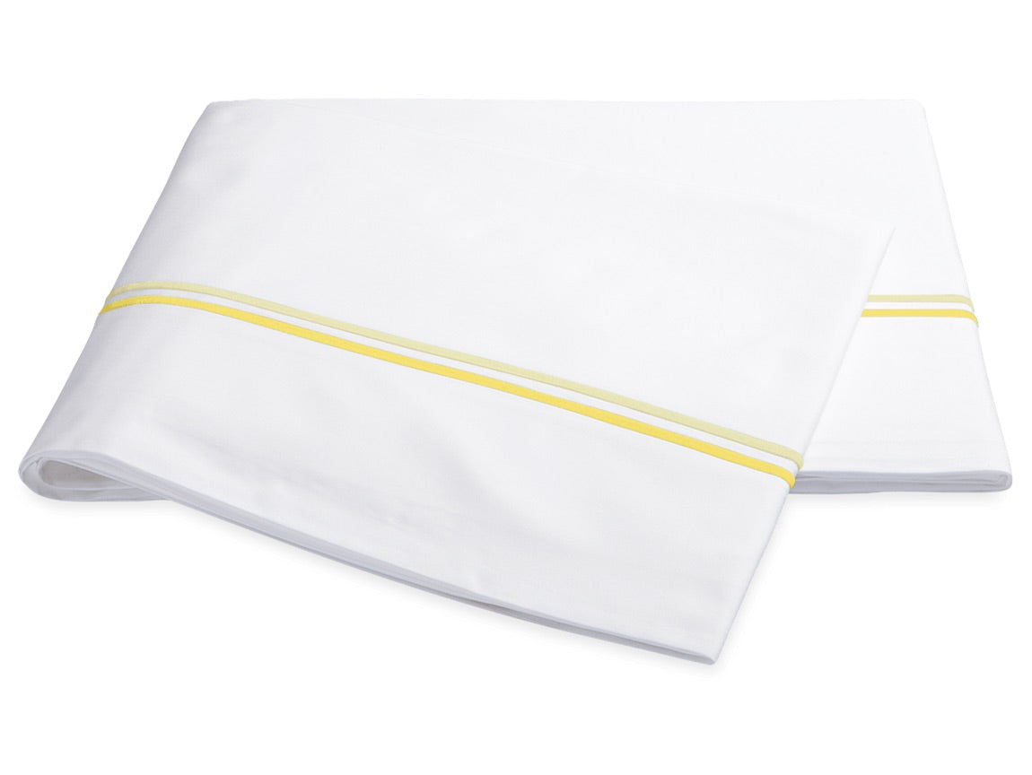 Two folded white sheets from the Essex Bedding Collection by Matouk, featuring a yellow stripe near the edge for a classic European hotel-inspired look, presented on a plain white background.