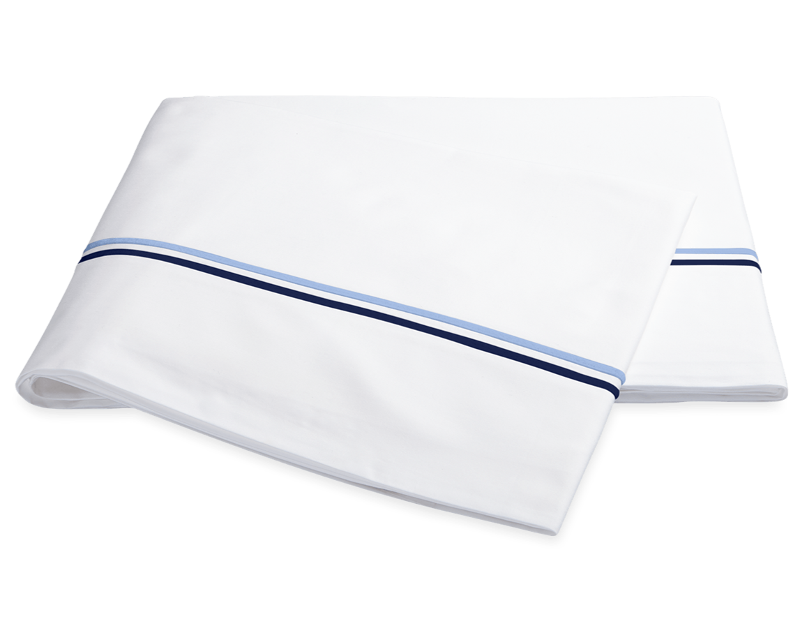 The Essex Bedding Collection in Navy by Matouk features folded white sheets with two parallel blue stripes near the edge, reminiscent of European hotel bedding, displayed on a plain white background.