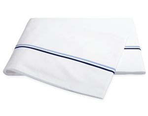 The Essex Bedding Collection in Navy by Matouk features folded white sheets with two parallel blue stripes near the edge, reminiscent of European hotel bedding, displayed on a plain white background.