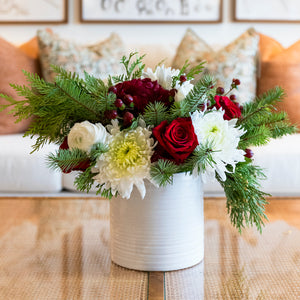 The Hive Floral Studio Live Evergreen Holiday Floral Arrangement features red roses, white chrysanthemums, greenery, and pine branches in a white ceramic vase, creating a festive accent for your living room.