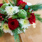 The Hive Floral Studio Live Evergreen Holiday Floral Arrangement features red roses, white chrysanthemums, green foliage, and red berries on a woven placemat—an ideal festive centerpiece for the holidays.