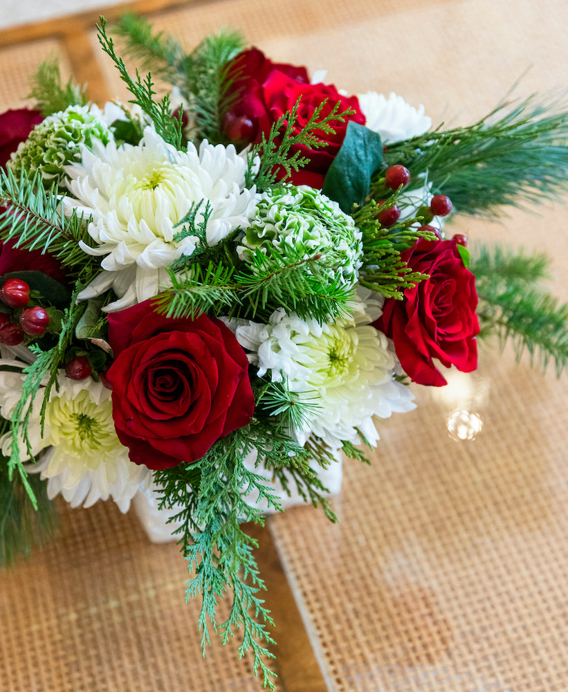 The Hive Floral Studio Live Evergreen Holiday Floral Arrangement features red roses, white chrysanthemums, green foliage, and red berries on a woven placemat—an ideal festive centerpiece for the holidays.