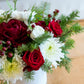 The Live Evergreen Holiday Floral Arrangement by Hive Floral Studio features red roses, white chrysanthemums, seasonal blooms, greenery, and red berries in a white vase on a woven surface.