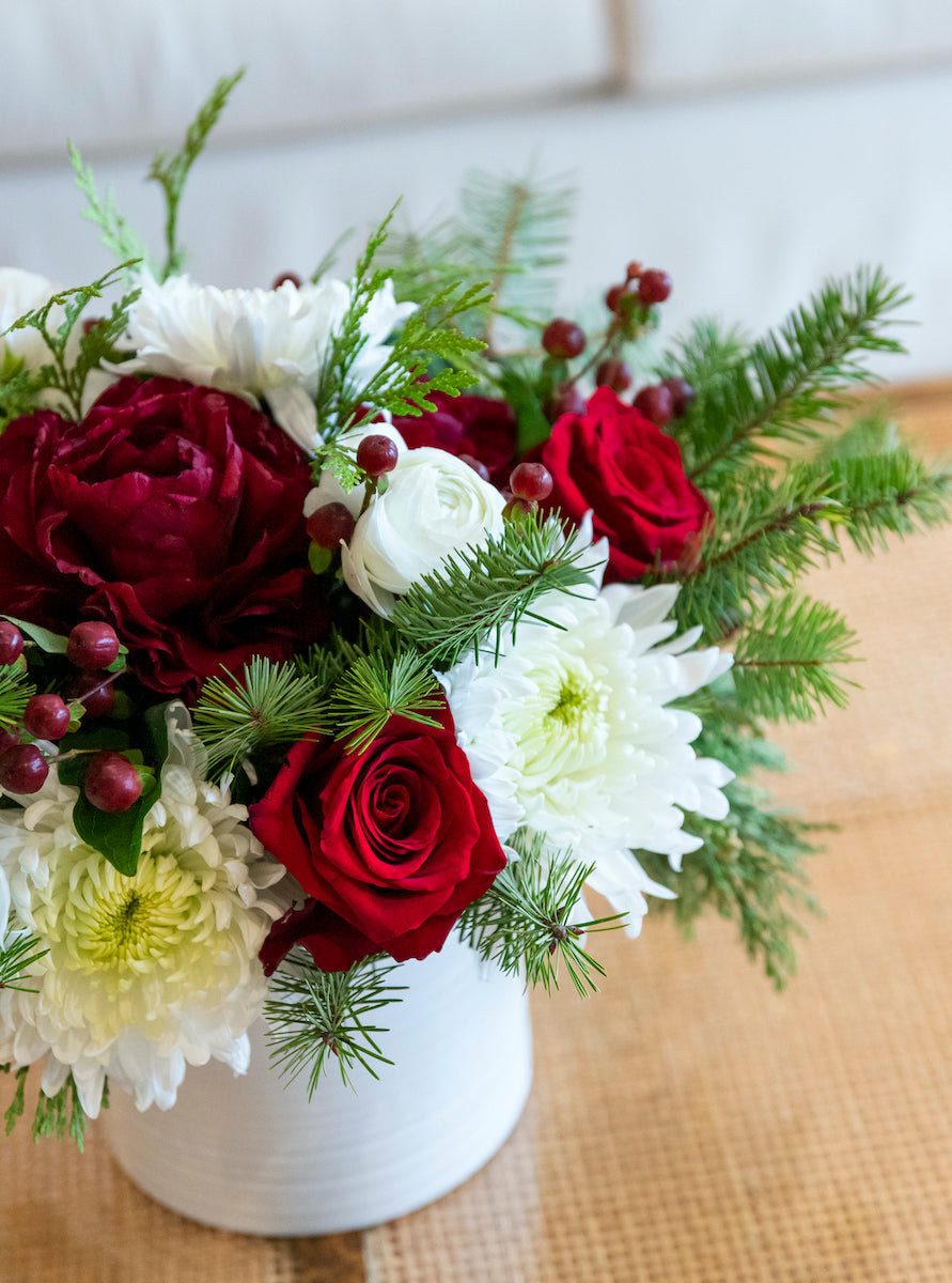 The Live Evergreen Holiday Floral Arrangement by Hive Floral Studio features red roses, white chrysanthemums, seasonal blooms, greenery, and red berries in a white vase on a woven surface.