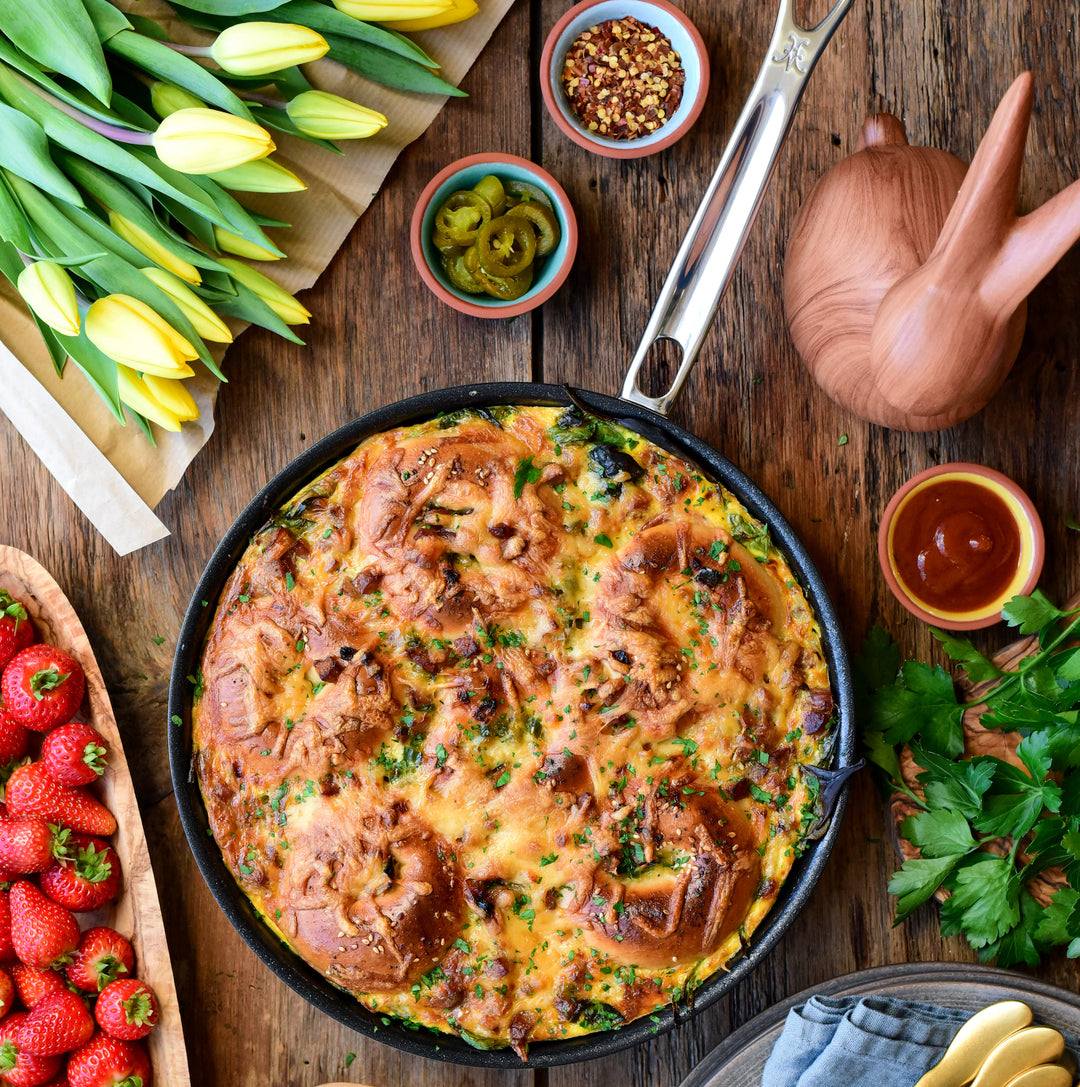 A baked egg casserole with bread and herbs is in a Hestan Thomas Keller Commercial Clad Stainless Steel TITUM® Nonstick Sauté Pan, 12.5", set on a wooden table with yellow tulips, strawberries, parsley, condiments, and cutlery.
