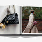 An open copy of Assouline's "Fashionphile: The Book of Iconic Bags" displays a close-up of a black quilted handbag and a woman in a beige coat with a pink quilted bag, highlighting the timeless appeal of luxury accessories.