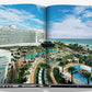 The open book displays an aerial view of a beachfront resort, featuring a large curved building reminiscent of the iconic Fontainebleau Hotel. Surrounded by pools and palm trees, luxurious lounges rest under a blue sky with fluffy clouds. Product: Fontainebleau by Assouline.