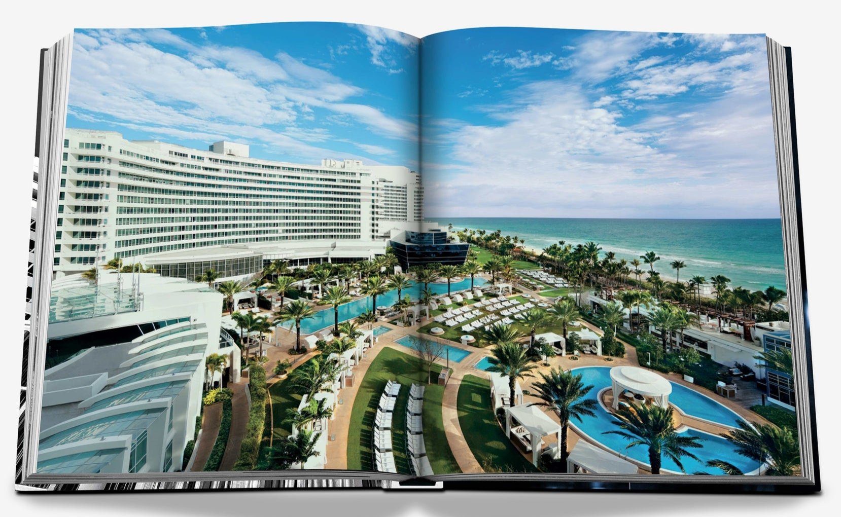 The open book displays an aerial view of a beachfront resort, featuring a large curved building reminiscent of the iconic Fontainebleau Hotel. Surrounded by pools and palm trees, luxurious lounges rest under a blue sky with fluffy clouds. Product: Fontainebleau by Assouline.