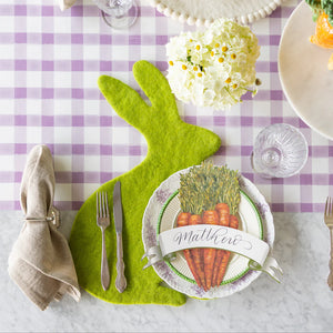 A whimsical table is set with Hester & Cook's Classic Moss Banner Table Accent, carrot-illustrated plate and name card, utensils, napkin, glass, a flower arrangement, all on a purple gingham tablecloth.