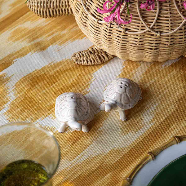 The Juliska Clever Creatures Turtle Salt and Pepper Set adds a touch of wisdom and charm on a patterned surface beside a wicker turtle basket, with an inviting green drink—ideal for any entertaining enthusiast.