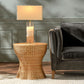 A Made Goods Nevis Side Table with a tapered silhouette and scalloped base, finished in wicker, supports a crystal lamp beside a dark gray velvet sofa; all are arranged on a light patterned rug against a paneled wall.