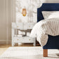 A Made Goods Wylder Nightstand with two drawers and shelf holds books beside a blue bed with striped bedding, topped by a wall-mounted sconce featuring a wicker shade.