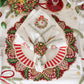 A Kim Seybert Christmas Trellis Napkin featuring an embroidered wreath, folded and secured with a beaded ring on a red and white striped dinner plate.