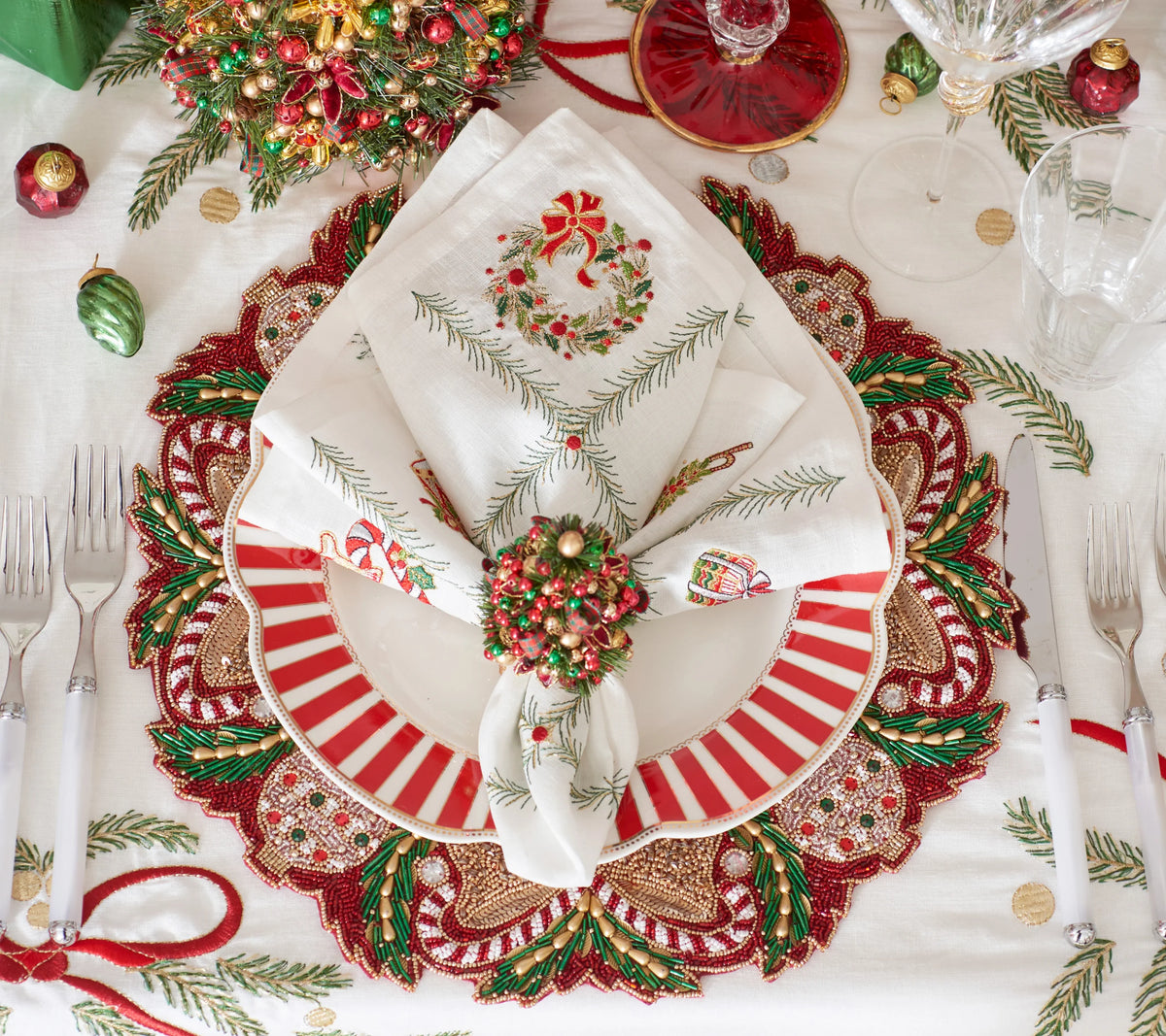 A Kim Seybert Christmas Trellis Napkin featuring an embroidered wreath, folded and secured with a beaded ring on a red and white striped dinner plate.