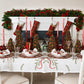 A Kim Seybert Merry and Bright Tablecloth embroidered with green pine branches and red ribbons, set with place settings, miniature decorated Christmas trees, and red taper candles.