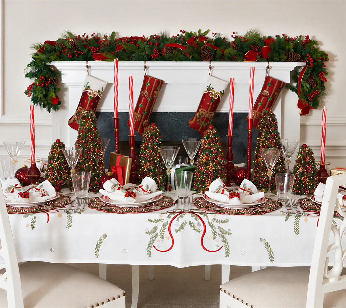 A Kim Seybert Merry and Bright Tablecloth embroidered with green pine branches and red ribbons, set with place settings, miniature decorated Christmas trees, and red taper candles.