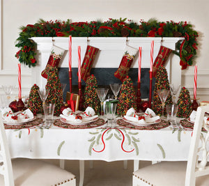 A Kim Seybert Merry and Bright Tablecloth embroidered with green pine branches and red ribbons, set with place settings, miniature decorated Christmas trees, and red taper candles.