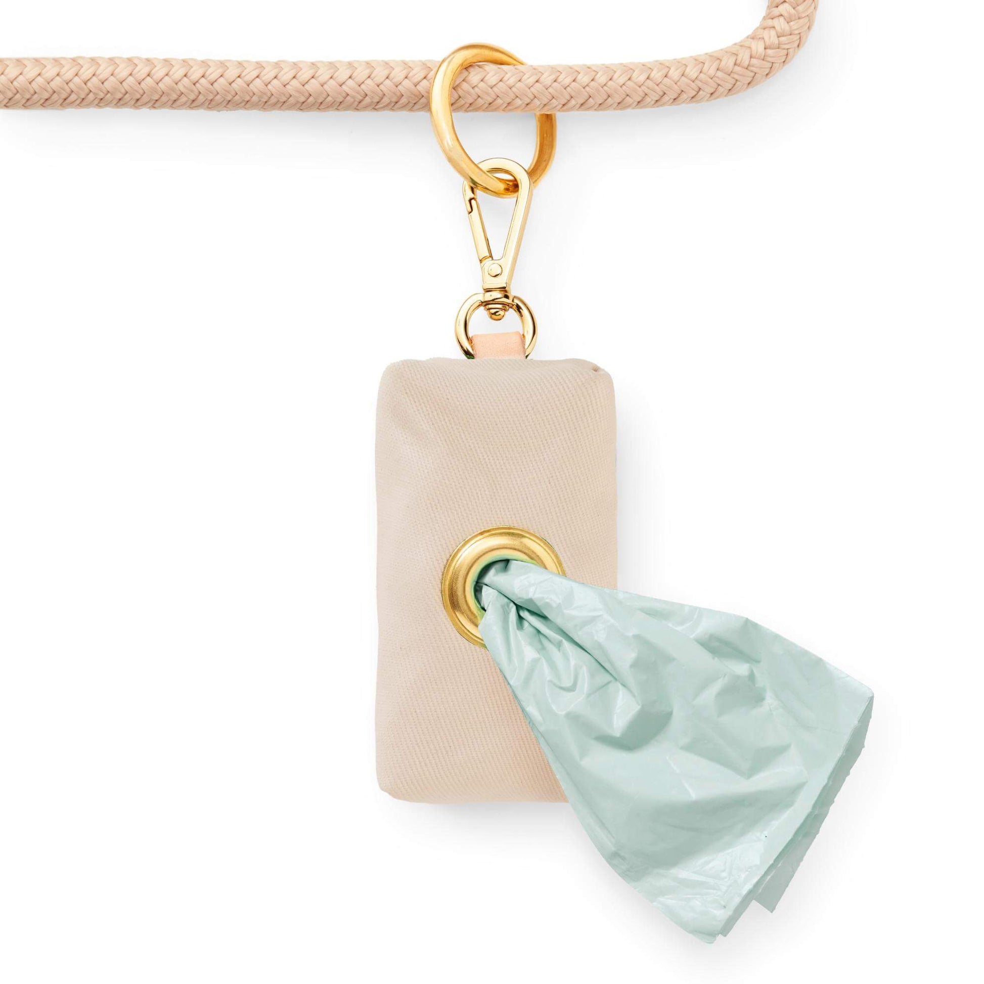 The Foggy Dog’s Waste Bag Dispenser in Flax, featuring gold hardware and attached to a light brown leash, dispenses green waste bags through a metal grommet—making your walks both convenient and stylish.