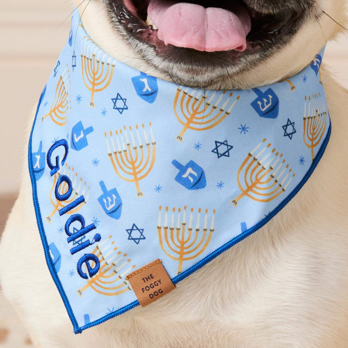The Festival of Lights Dog Bandana is a light blue bandana featuring a pattern of gold menorahs, blue dreidels, and Stars of David, personalized with the name “Goldie” embroidered in blue script.