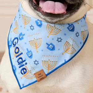 The Festival of Lights Dog Bandana is a light blue bandana featuring a pattern of gold menorahs, blue dreidels, and Stars of David, personalized with the name “Goldie” embroidered in blue script.