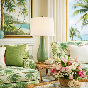 A living room with tropical decor combines modern style, featuring palm leaf sofas, the Visual Comfort Form Table Lamp in Shellish Kiwi, a beach painting, and a floral arrangement atop a sculptural wicker table.