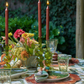 An outdoor dining table set features green plates, hand-blown glassware, a floral centerpiece, and red candles in the Iris Tall Candle Holder Set by Kim Seybert, all surrounded by lush greenery.