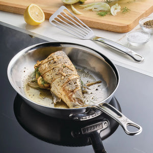 A whole fish tied with herbs and lemon slices cooks in the Hestan Provisions Oval Fish Pan on the stovetop, with a spatula, sliced fennel, lemon, salt, and pepper nearby—perfect for chef-style preparations.
