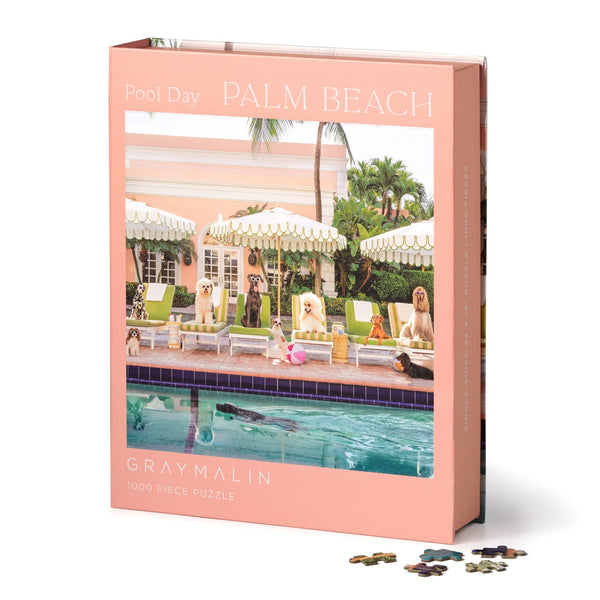 The Gray Malin 1000 Piece Puzzle by Chronicle Books features a Palm Beach poolside scene with dogs lounging and palm trees in a pink and peach palette, evoking The Colony Hotel.