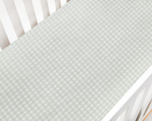 A close-up of the Gooselings Green Picnic Gingham Crib Sheet, OEKO-TEX certified in light green and white, covers a crib mattress inside a white crib.