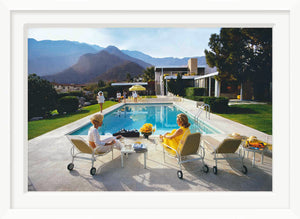 Two individuals relax on poolside loungers beside a swimming pool, evoking Slim Aarons' iconic Palm Springs allure. Between them is a table with drinks and flowers, backed by mountains and a modern Richard Neutra-style house in this Getty Images photo, "Poolside Glamour," from Soicher Marin.