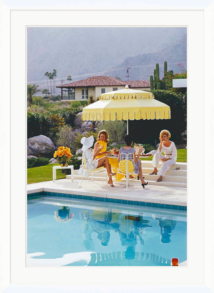 Three women sit by a pool, chatting under a yellow umbrella reminiscent of Slim Aarons style. One holds a drink with the iconic Kaufmann Desert House and Palm Springs landscape as their backdrop, captured in Soicher Marin's "Getty Images, 'Nelda and Friends'," Slim Aarons, January 1, 1970.