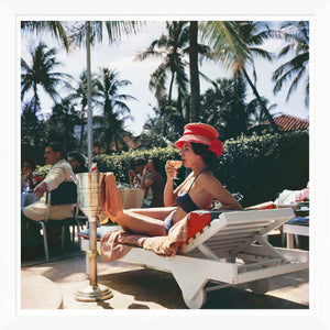 A woman in a black bikini and red hat relaxes poolside, drink in hand, evoking Slim Aarons' iconic style in Getty Images' “Leisure and Fashion” (1961) by Soicher Marin—complete with diners and palm trees in the background.