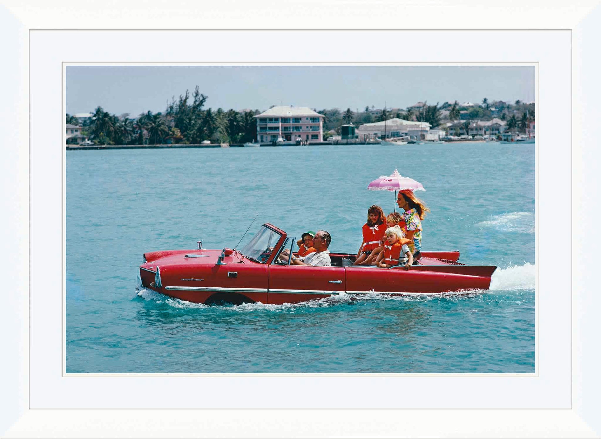 Getty Images’ “Sea Drive” by Slim Aarons, offered by Soicher Marin, shows a red amphibious car with six people gliding on water amid sunny buildings and trees, evoking luxury and classic style from 1967.