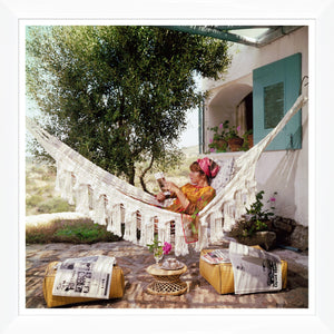 A woman reads in a fringed hammock outside a white house with green shutters, evoking the iconic style of Slim Aarons as seen in Getty Images’ "Bettina Grazini," 1964, by Soicher Marin.