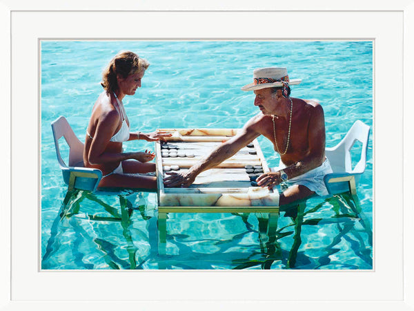 Two people sit in clear shallow water playing backgammon on a floating board, mirroring Slim Aarons' style. With the man in a hat and sunglasses, both enjoying swimwear, it's a timeless leisure scene reminiscent of Slim Aarons' "Keep Your Cool" from 1978 by Soicher Marin via Getty Images.