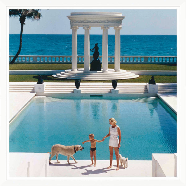 A woman and child are by a large pool with two dogs, capturing the timeless charm of "Nice Pool," a Slim Aarons photo from January 1, 1955. A white pavilion and the ocean in the background add to its Villa Artemis-like allure, available from Soicher Marin.