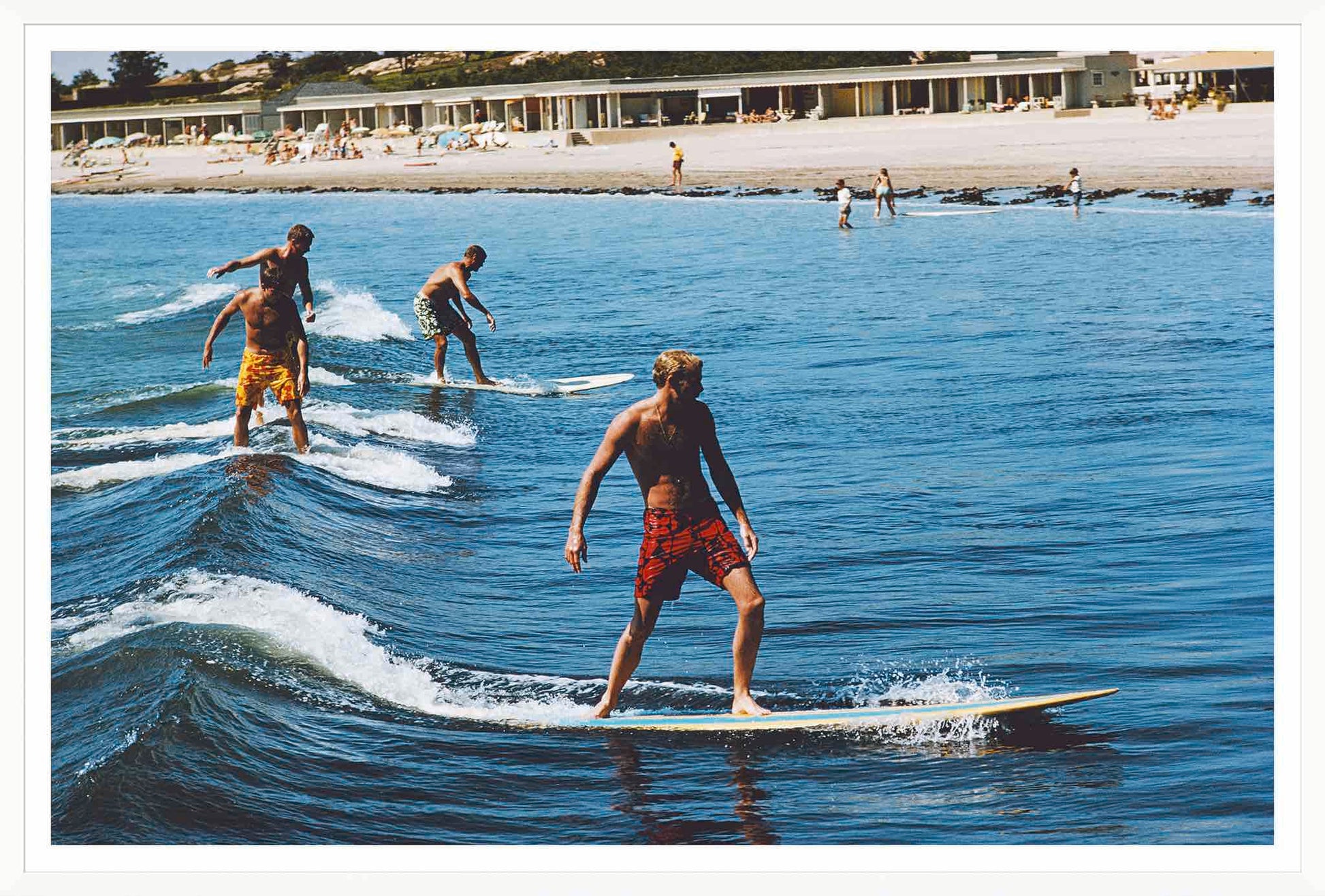 Four people surf small waves near a sandy beach, sunbathers and buildings behind them—a scene from "Surfing Brothers" by Slim Aarons (Getty Images, 1965), available from Soicher Marin.